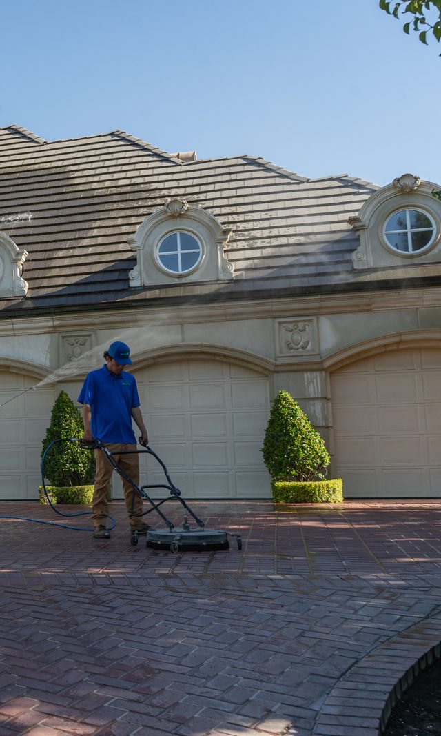 Valley Wash Pros technician running a surface cleaner on a brick driveway in front of a large home
